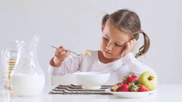 Portrait of sad small girl doesn't want to eat cornflakes with milk for breakfast. She is looking on her food with spoon sitting at table on white background.