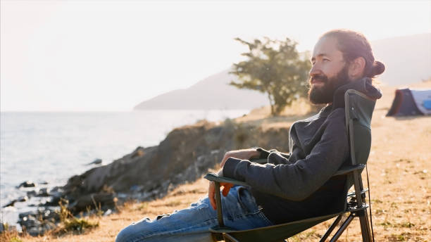 Young man sits on chair at campsite on lake shore stock photo