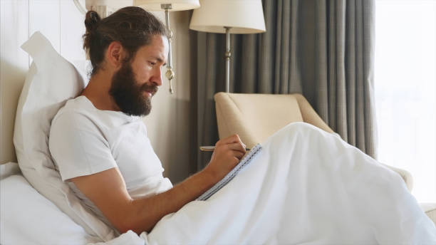 Young bearded man writing notes to diary after awake in bedroom at home stock photo
