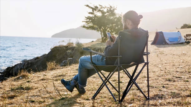 istock Young man sitting on folder chair and using smart phone at camping at lake shore 1184545780