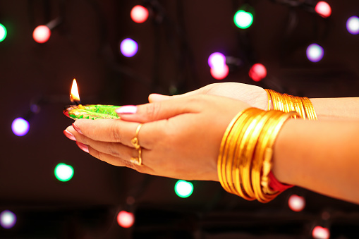Woman hands with bangles holding Diwali diya clay lamps in close up view on dark bokeh background
