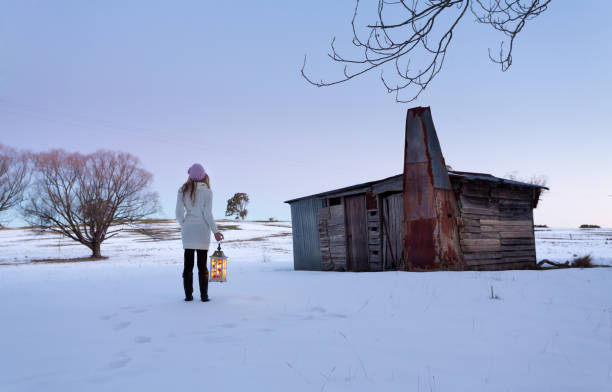 Woman with lantern standing in snow by old rustic shed or hut