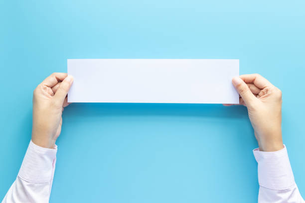 istock close up hands holding empty white blank horizontal paper for flyer or invitation mock up isolated on a blue background. 1184563983