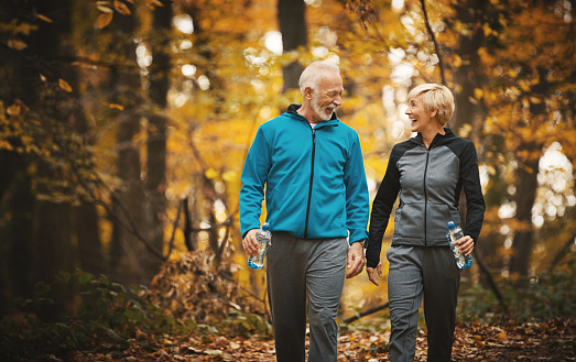 Closeup front view of a senior couple walking in a forest and having fun. They are enjoying these beautiful fall colors while stepping over dry leaves and sipping water from water bottles.
