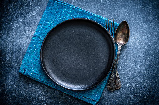 Top view of a black plate with a spoon and fork at the right side shot against a textured bluish toned background. A blue textile napkin is under the plate. Predominant colors are blue and black. Low key XXXL 42Mp studio photo taken with Sony A7rii and Sony FE 90mm f2.8 macro G OSS lens