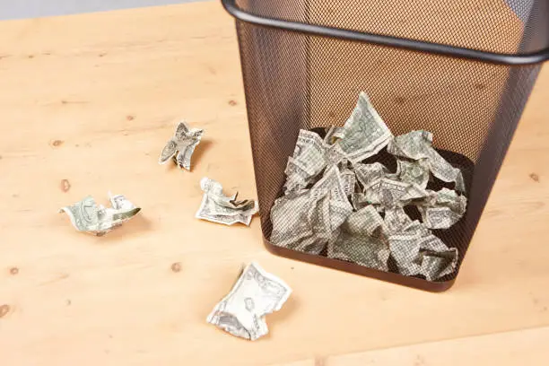 Many One dollar bills have been thrown into a trash can with more on the wooden floor next to it.