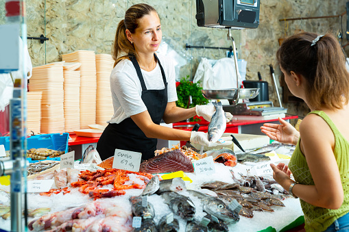 Polite female worker of fish store offering fresh sea bass to woman customer