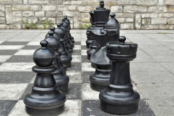 istock Black plastic big chess pieces are weathered but neatly lined up on a weathered chessboard outside on paving stones 1184600820