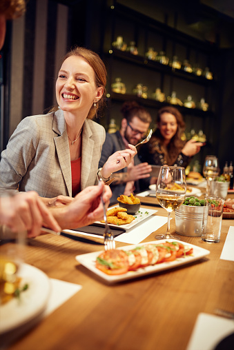 Charming smiling caucasian blonde sitting in restaurant, eating dinner and chatting with friend. In background are friends.