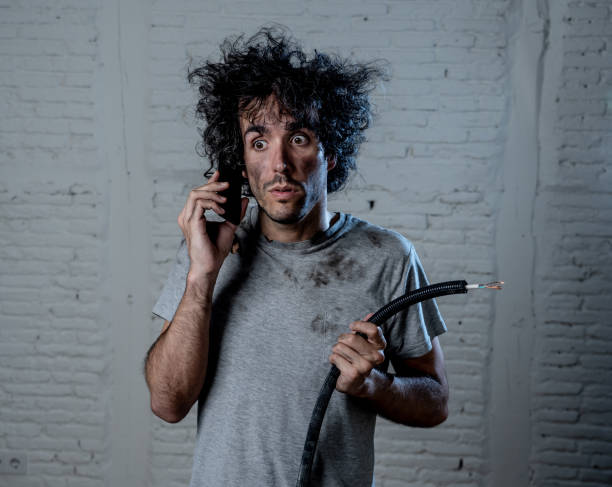 Portrait of young man holding electrical cable after domestic accident with dirty burnt funny face expression calling desperate with mobile phone asking for help. Electricity repairs and DIY concept. stock photo