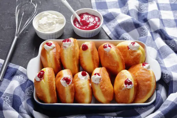 close-up of Berliner Pfannkuchen, German Donuts with Raspberry Jam and cream fillings in a baking dish on a concrete table with jam and cream in a bowls, view from above