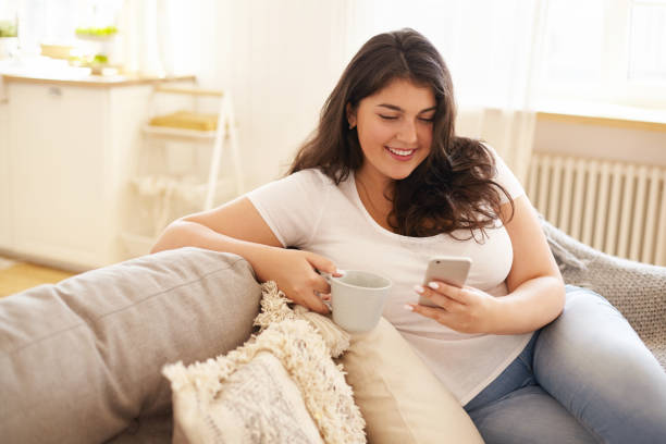 tecnologia, comunicazione e concetto di svago. bella ragazza con guance paffuto rilassanti a casa con gadget elettronico. giovane donna bruna in sovrappeso che sms sullo smartphone e prendere un caffè - sovrappeso foto e immagini stock