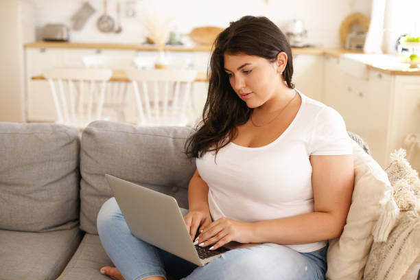 adorabile ragazza studentessa plus size con tastiera per capelli neri sciolti su laptop seduta comodamente sul divano in salotto, sorridente, messaggia amici online, utilizzando la connessione internet wireless ad alta velocità - comfortably foto e immagini stock