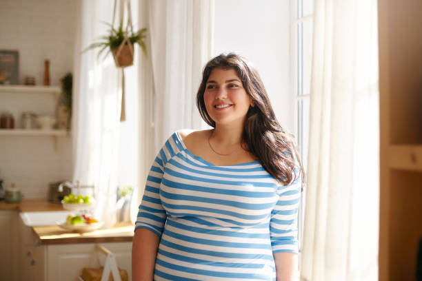 beauty, bodypositivity, people and lifestyle concept. indoor shot of gorgeous brunette latin girl posing by window in kitchen dressed in xxl blue and white striped shirt, having joyful beaming smile - women smiling body cheerful imagens e fotografias de stock