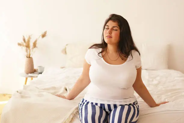 Photo of Beautiful relaxed young woman with curvy body and black hair keeping her eyes closed. Brunette cute plus size girl enjoying nice day, relaxing in bedroom, wearing striped trousers and white t-shirt
