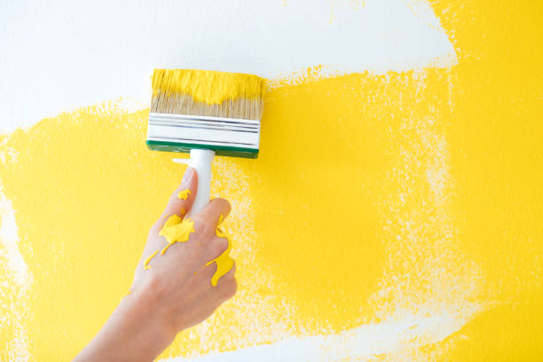 a mão fêmea pinta uma parede branca com uma escova e uma tinua amarela, gotas da pintura fresca líquida escorre abaixo da mão - house painter paint roller yellow painting - fotografias e filmes do acervo