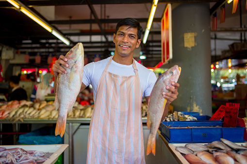 A fish stall owner holding fishes and smiling in front the camera at his stall
