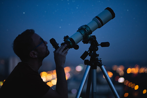 Astronomer with a telescope watching at the stars and Moon with blurred city lights in the background.