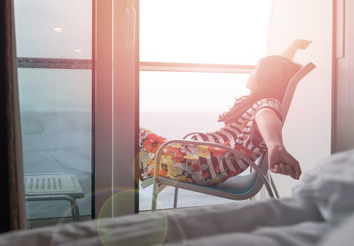 Relax business woman lifestyle at cruise ship sitting on chair in balcony looking out to beautiful sea landscape nature with sunlight effect during year end holiday vacation trip.