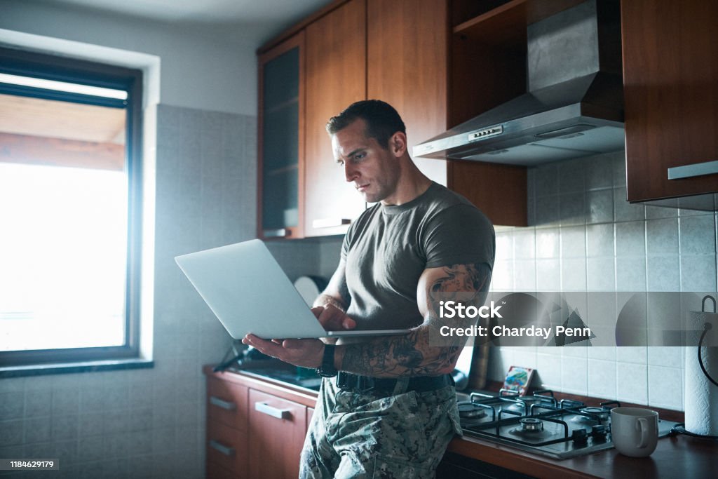 Beat the army blues by staying connected Shot of a handsome young soldier standing in the kitchen and using a laptop Military Stock Photo