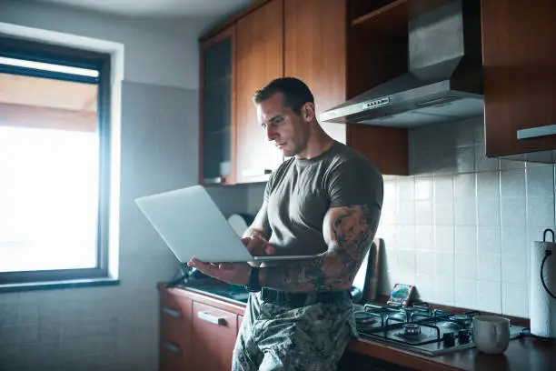 Shot of a handsome young soldier standing in the kitchen and using a laptop
