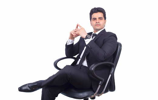 Handsome young businessman sitting on chair. Confident male professional is against white background. He is wearing suit.