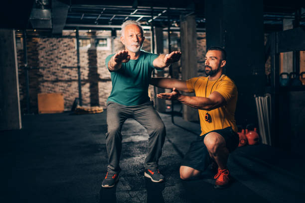 istock Active, white-haired elderly man doing squats with the assistance of his mentor in a   gym 1184649210