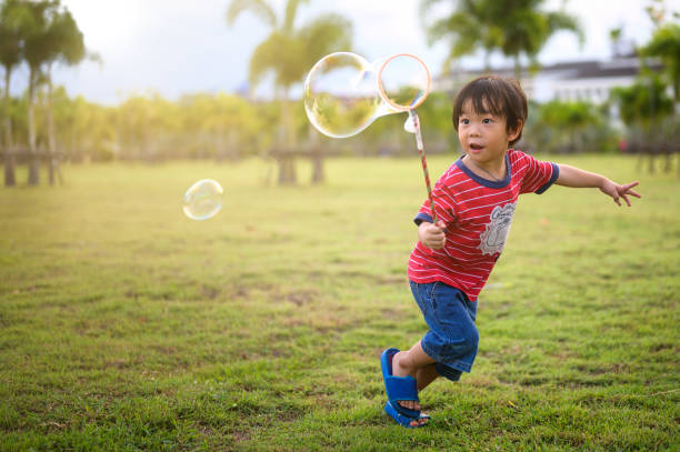 asiático feliz niño soplando y jugando jabón de varita burbuja en el jardín - bubble wand bubble child playful fotografías e imágenes de stock