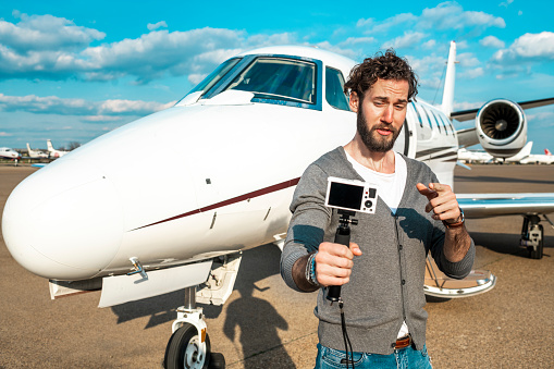 Male vlogger making a video on a small camera next to the nose cone of a private airplane parked on an airport taxiway.