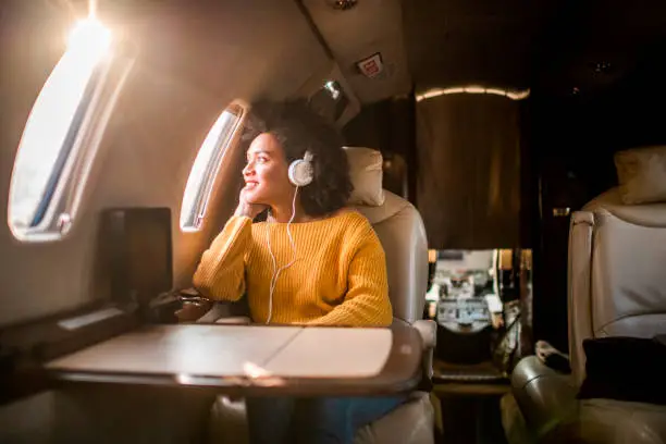 Young fashionable woman sitting on a private airplane and looking through a window while listening to music through headphones.