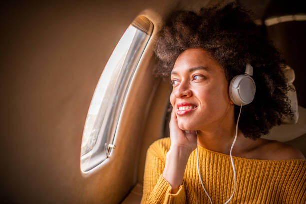 jeune femme chic s'asseyant sur un avion privé et regardant par une fenêtre tout en écoutant la musique par des écouteurs - status symbol audio photos et images de collection