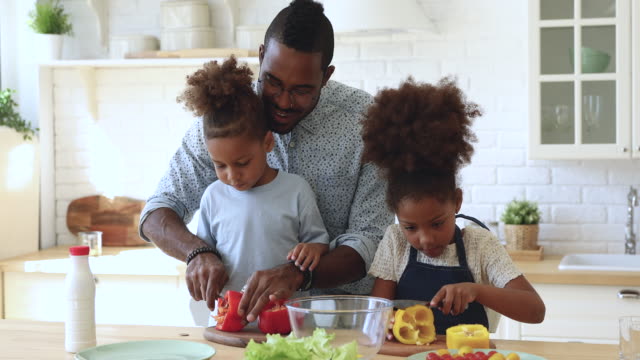 African father teaching son daughter cutting vegetable salad in kitchen
