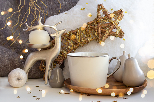 Cup of coffee tea on Christmas composition with candle, deer, fur pillow, and Xmas baubles on New Year background with bright lights and gold stars. Set of warmth and comfort.