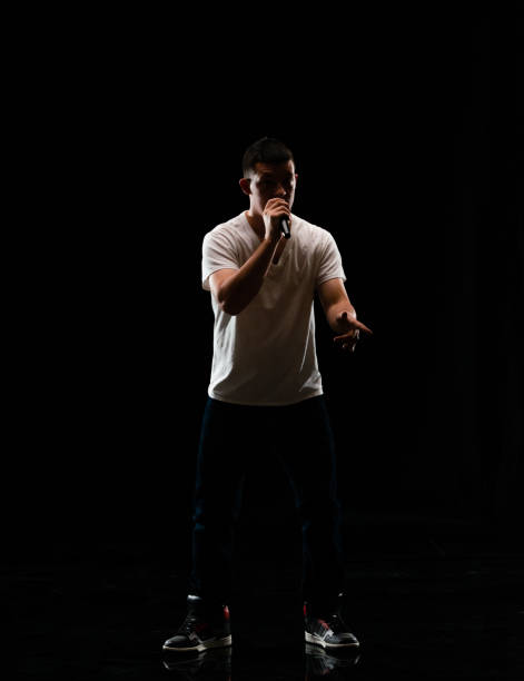 Full length / one man only / one person / front view of 20-29 years old adult handsome people caucasian male / young men singer / musician standing / dancing in front of black background wearing canvas shoe / jeans / pants / t-shirt / shirt who is singing / happy and holding microphone / black color / back lit / hip-hop longitud completa / un hombre solamente / una persona / vista frontal de 20-29 años de edad adulto guapo personas caucásicas hombres / hombres jóvenes cantante / músico de pie / bailando frente al fondo negro usando zapato de lona / jeans / pantalones