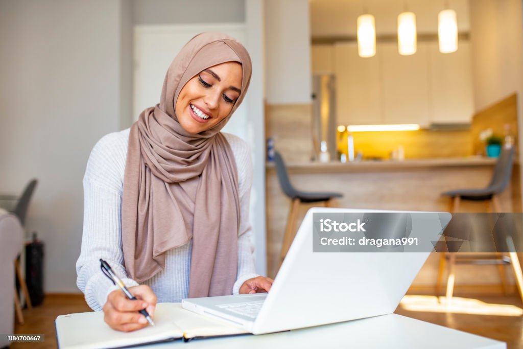 Essential for getting her tasks done Attractive female Arabic working on laptop computer and paperwork's on desk. Arabian Businesswoman working at home. Dedication and technology. Essential for getting her tasks done Women Stock Photo