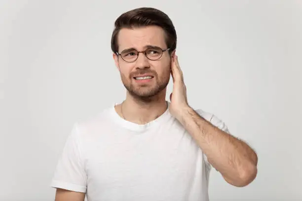 Unhealthy young man in eyeglasses touching ear, suffering from sudden throbbing ear ache, looking aside, head shot. Upset millennial guy feeling unwell, isolated on grey white studio background.