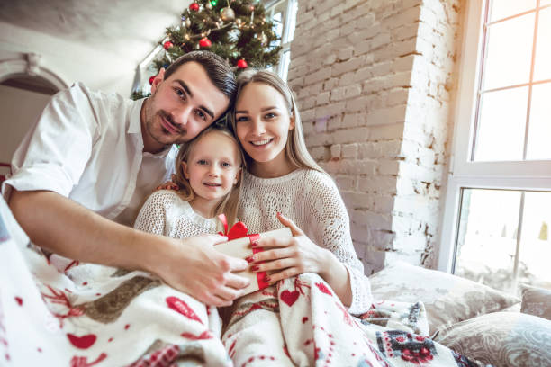 frohe weihnachten und ein gutes neues jahr! glückliche familie mit geschenk-box sitzt auf dem bett zu hause in der nähe von weihnachtsbaum. - gift new year couple men stock-fotos und bilder