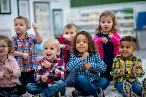 A group of multi-ethnic preschool students sit on a classroom rug.  They are singing, doing actions, clapping and having fun during a sing-along time.  Each is wearing casual clothing in bright colors.