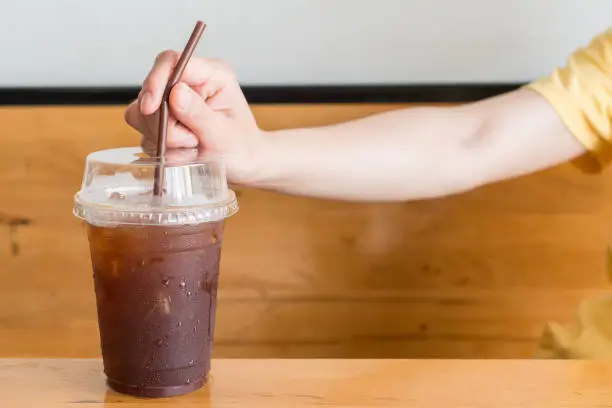 Photo of Close-up of woman hand with a plastic cup of iced Americano on the wooden table.