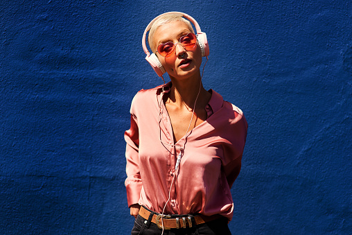 Cropped portrait of an attractive young woman standing against a blue wall and listening to music through headphones