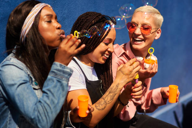 Cropped shot of three attractive young women sitting against a blue wall together and bonding by blowing bubbles