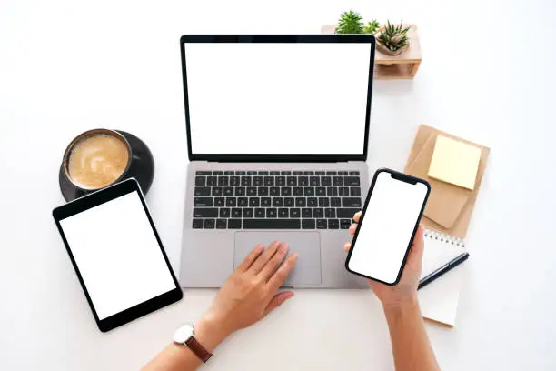 Photo of hands holding a blank white screen mobile phone with laptop computer and tablet pc on the table in office