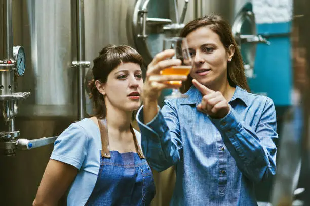 Close-up of Hispanic women in mid 30s working in craft beer brewery and checking sample for quality.