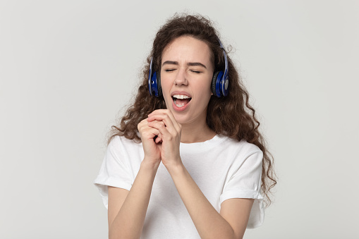 Head shot portrait overjoyed happy young woman wearing bluetooth overhead headphones, listening to popular music, pretending singing favorite song in microphone isolated on grey studio background.