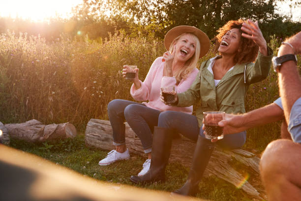 grupo de amigos maduros sentados alrededor del fuego mientras beben y cantan canciones en el camping al aire libre - outdoors drinking women friendship fotografías e imágenes de stock
