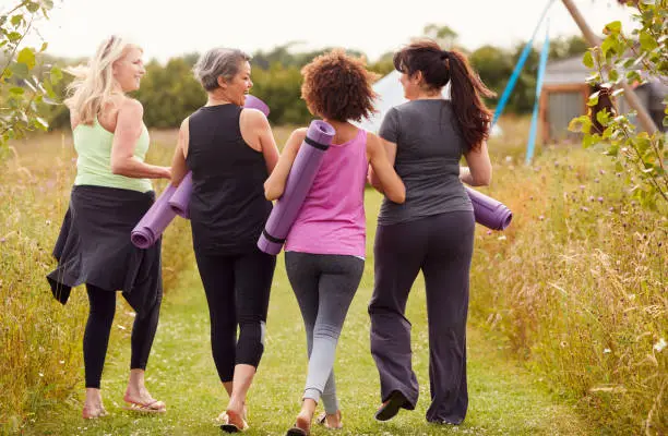 Rear View Of Mature Female Friends On Outdoor Yoga Retreat Walking Along Path Through Campsite