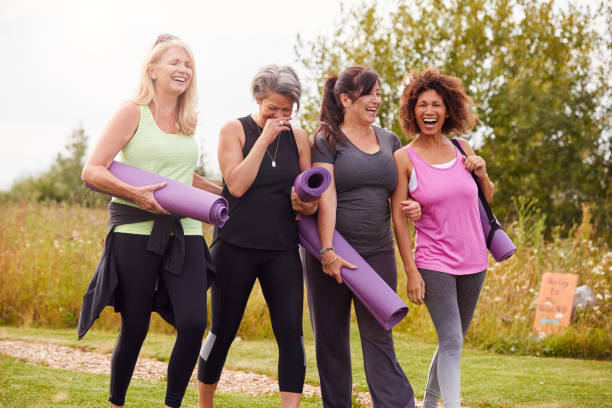 Group Of Mature Female Friends On Outdoor Yoga Retreat Walking Along Path Through Campsite Group Of Mature Female Friends On Outdoor Yoga Retreat Walking Along Path Through Campsite exercise mat photos stock pictures, royalty-free photos & images