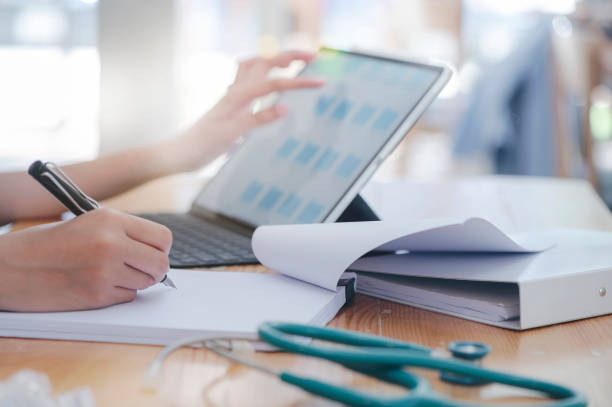 istock Closeup female doctor hand writing on paperwork with pen while working with tablet at table in hospital. 1184840865