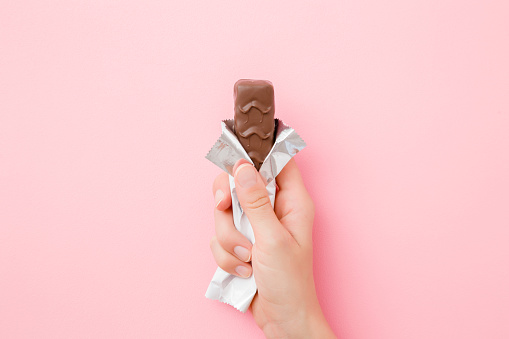 Young woman hand holding chocolate bar on pastel pink table. Opened pack. Sweet snack. Closeup. Top view.
