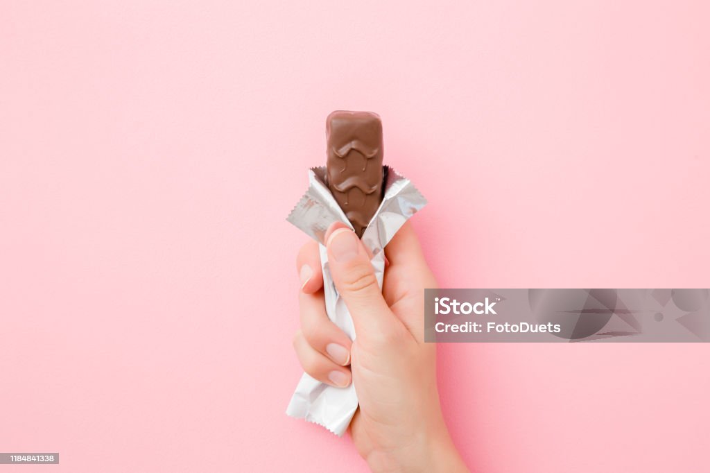 Young woman hand holding chocolate bar on pastel pink table. Opened pack. Sweet snack. Closeup. Top view. Chocolate Bar Stock Photo